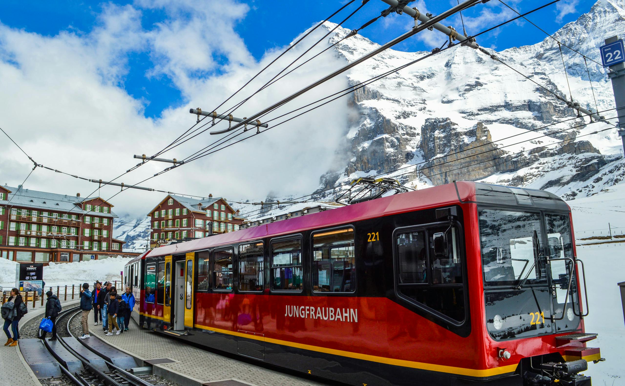 From below of contemporary train on railway near group of anonymous travelers on platform behind snowy mountain and buildings in winter